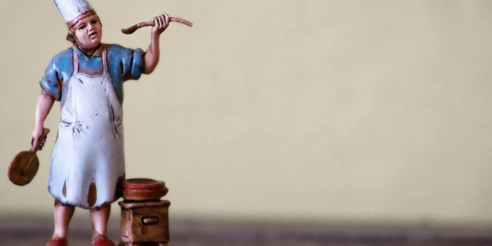 Una statuetta di un cuoco con cappello e grembiule bianco si trova accanto a una pentola su un fornello, con in mano un cucchiaio di legno e un mattarello: perfetto per chi sogna di assumere un cuoco personale Varese per creare un menù su misura.