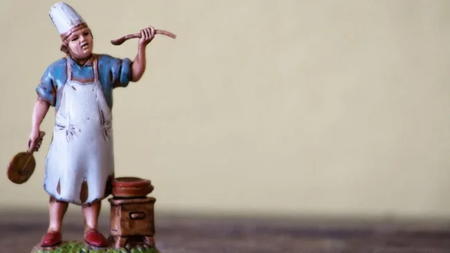 Una statuetta di un cuoco con cappello e grembiule bianco si trova accanto a una pentola su un fornello, con in mano un cucchiaio di legno e un mattarello: perfetto per chi sogna di assumere un cuoco personale Varese per creare un menù su misura.