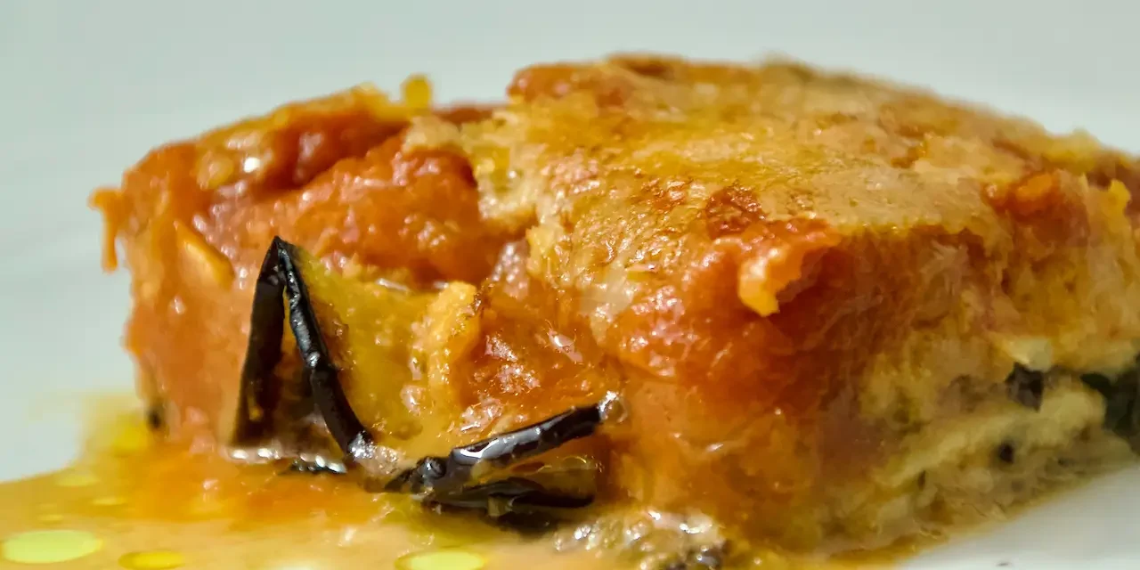 Primo piano di una fetta di parmigiana di melanzane con strati di salsa di pomodoro, formaggio fuso e melanzane al forno su un piatto bianco, presentata da Giorgio di Cucino di Te, chef a domicilio a Varese.