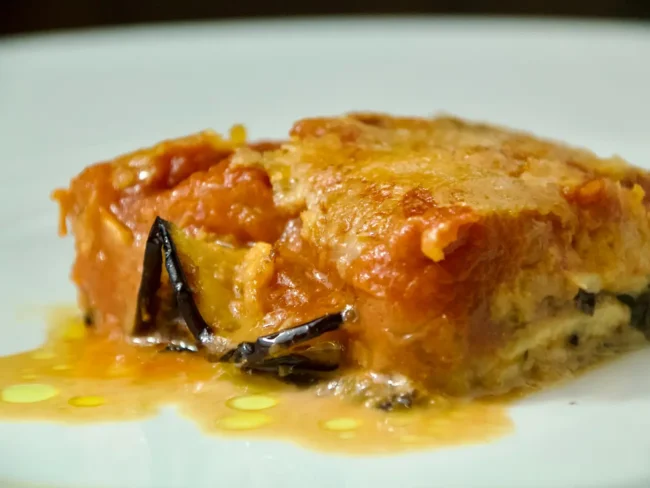 Primo piano di una fetta di parmigiana di melanzane con strati di salsa di pomodoro, formaggio fuso e melanzane al forno su un piatto bianco, presentata da Giorgio di Cucino di Te, chef a domicilio a Varese.